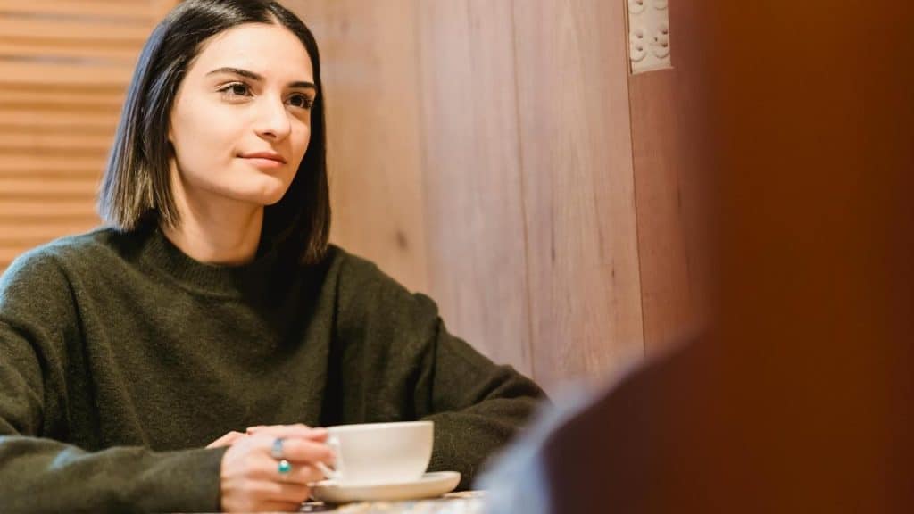 A woman holding a cup while sitting at a table and listening attentively.