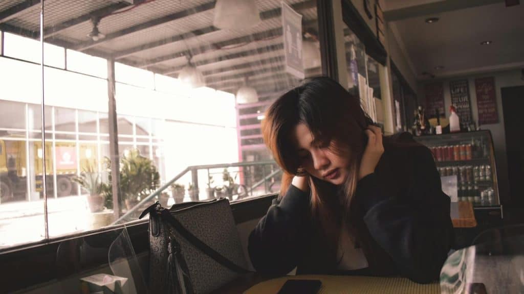 A woman sitting alone at a café table, looking down pensively beside her bag and phone.