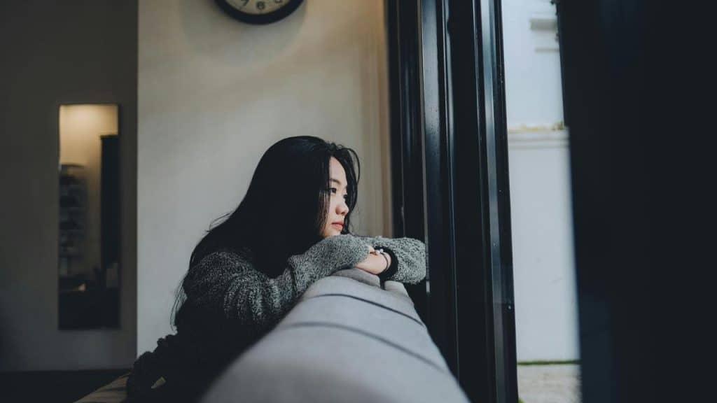 A woman leaning on a windowsill, gazing outside thoughtfully in a softly lit room.