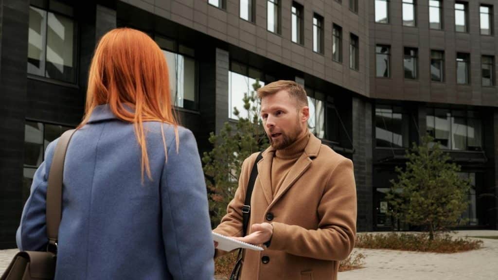 A man speaking to a woman outside a modern building, holding a notebook and gesturing.