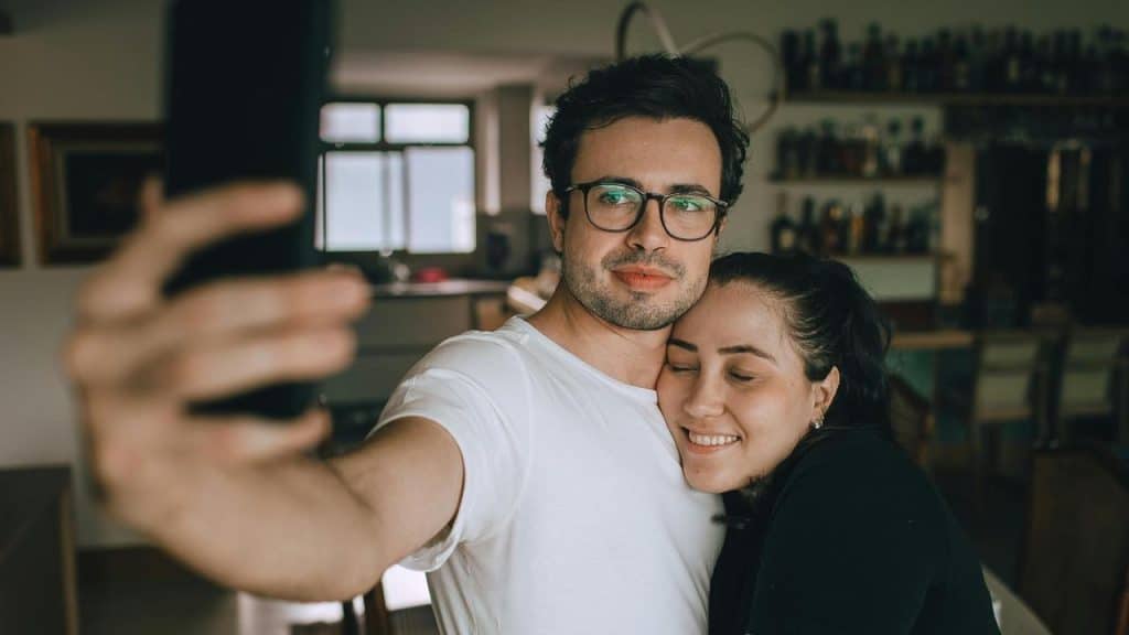 A couple taking a selfie indoors, smiling and leaning close together.