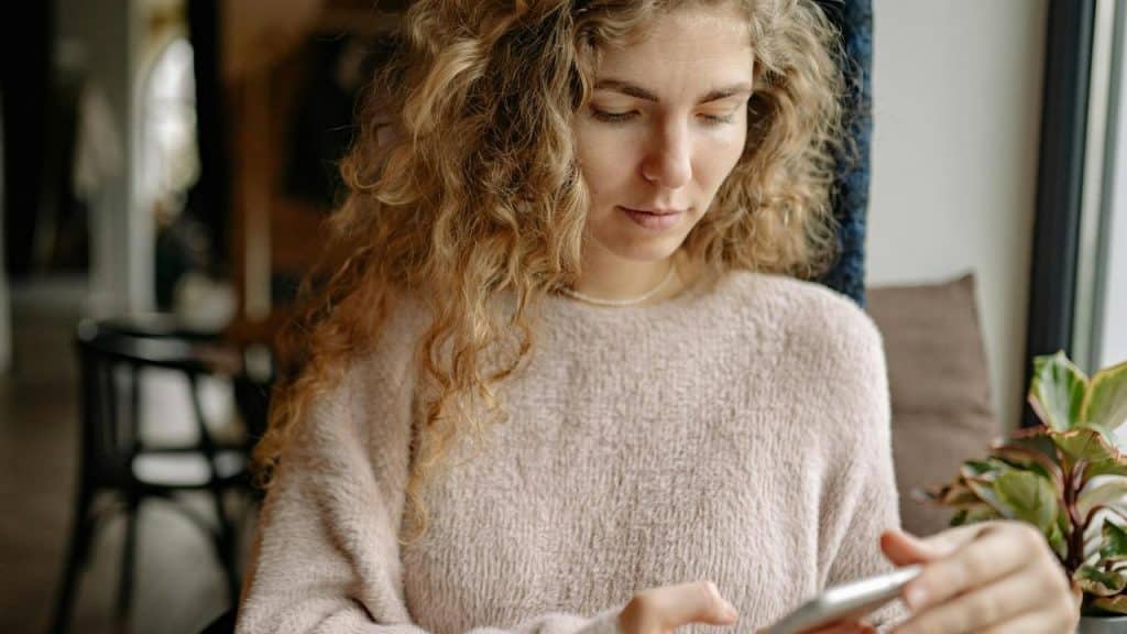 A woman sitting indoors, looking down at her phone with a focused expression.