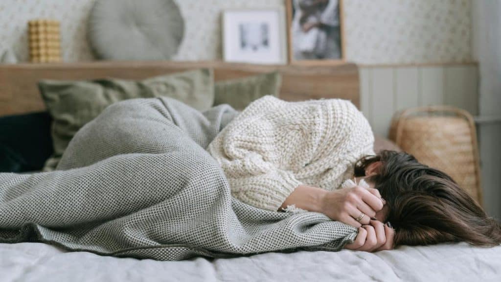 A person curled up on a bed under a blanket, appearing unwell or resting.