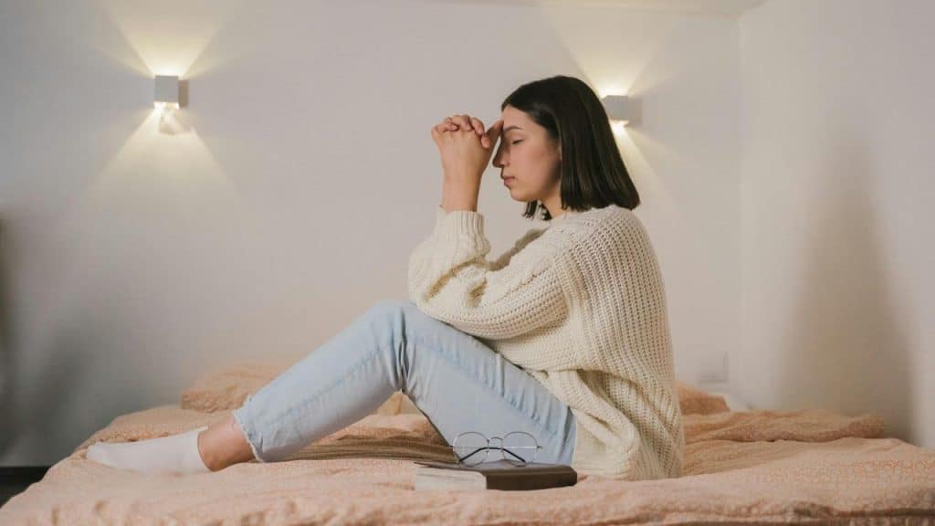 A woman sitting on a bed with eyes closed, holding her hands to her forehead.