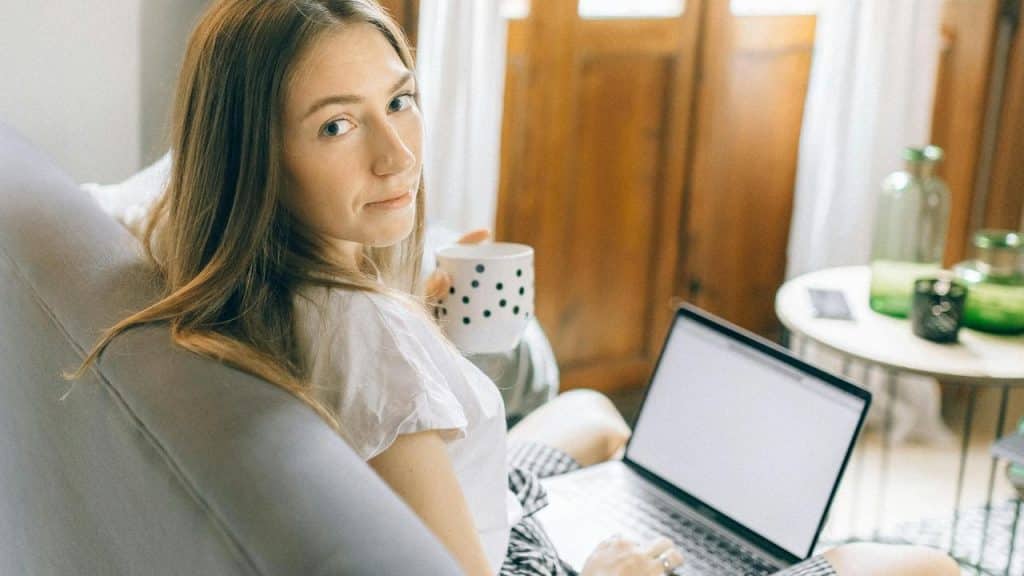 A woman sitting with a laptop, holding a mug and looking over her shoulder.