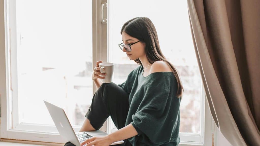 A woman sitting by a window with a laptop, holding a mug and looking down.
