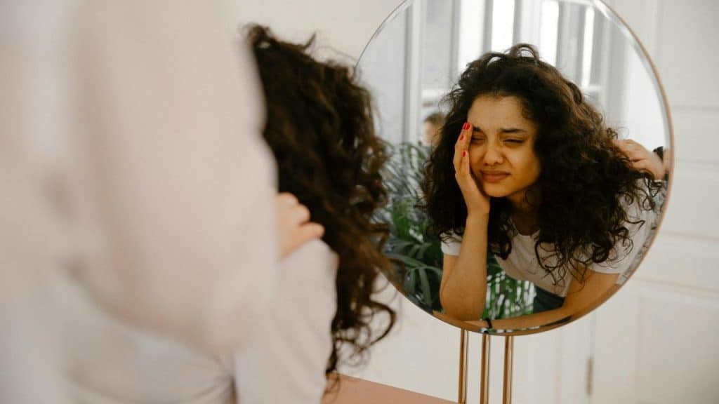 A woman looking in a mirror, touching her face with a pained expression.