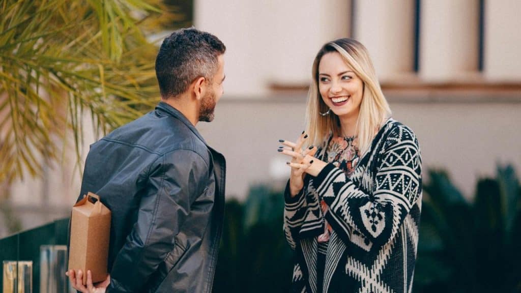 A smiling couple talking outdoors as one hides a gift behind his back