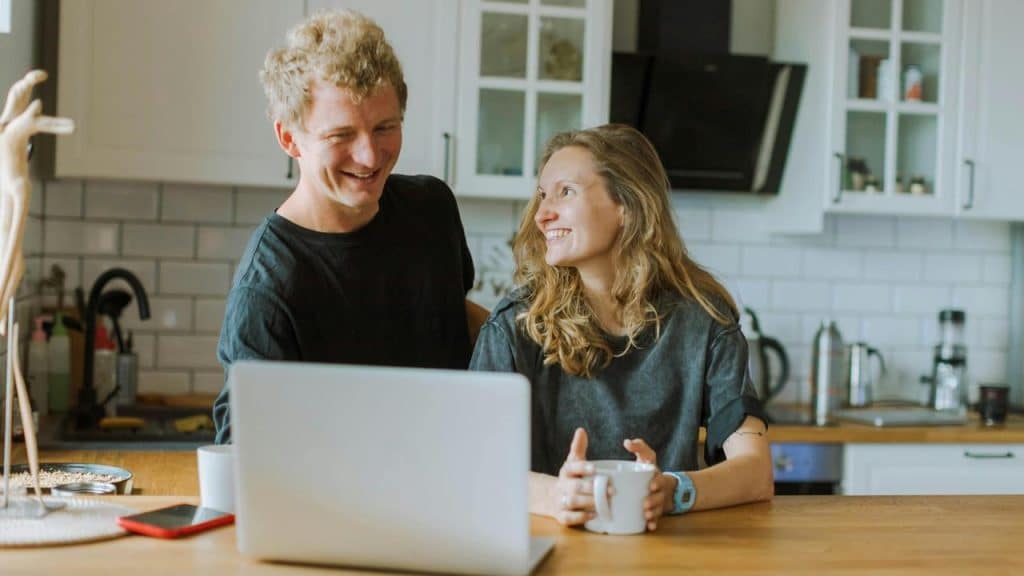 A smiling couple sitting at a kitchen table with a laptop and mugs