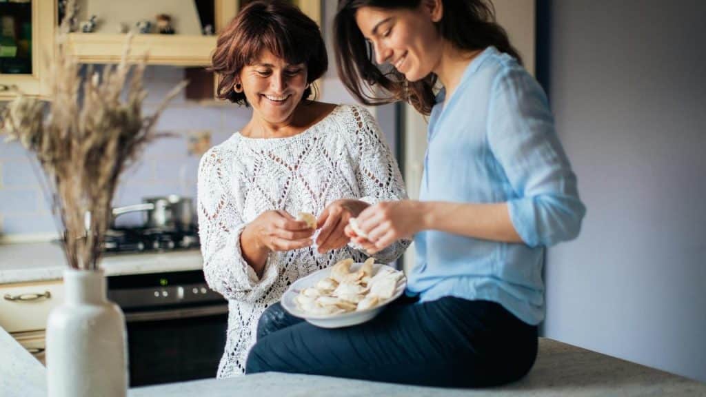 A smiling older woman and younger woman preparing food together in a kitchen.