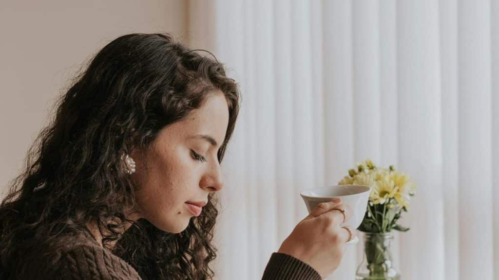 A woman holding a cup, looking down thoughtfully near a window.