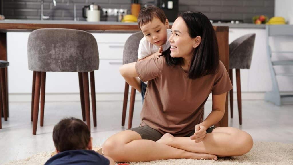 A woman sitting on the floor smiling while a child climbs on her back and another plays nearby.