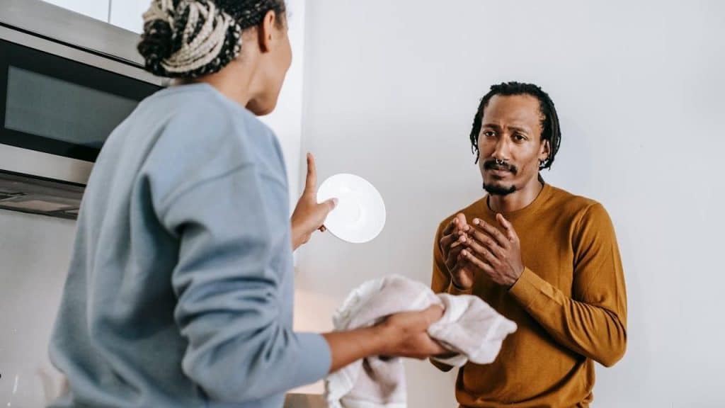A woman is holding a plate and towel, gesturing while speaking to a man who looks distressed
