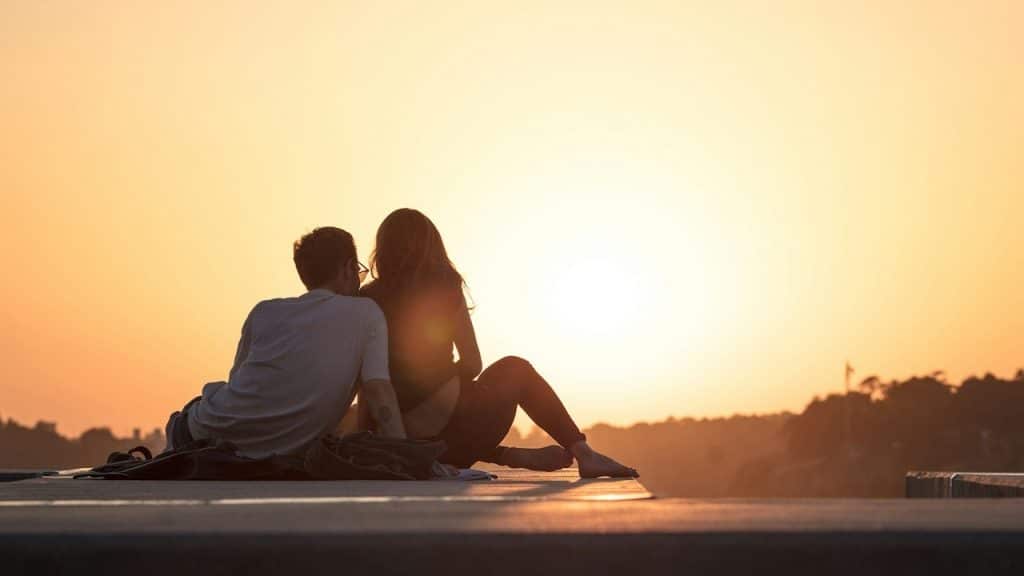A man and woman sitting on the ground and watching a sunset.