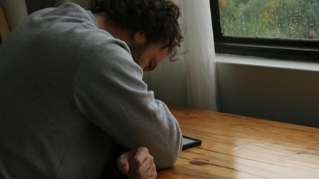 A person leaning over a table, looking down at a phone near a rain-streaked window.