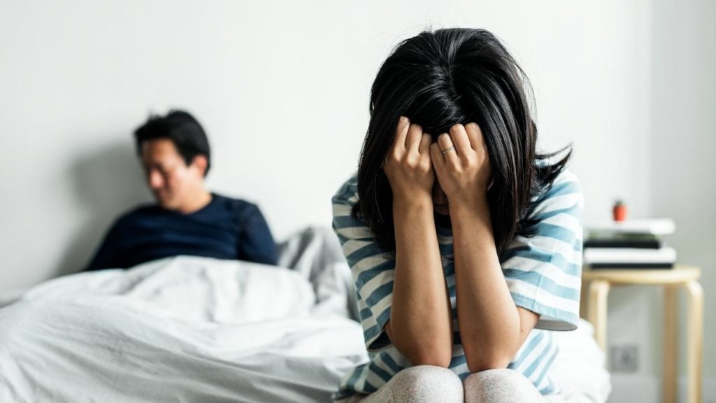 A distraught woman is sitting on a bed with her head in her hands, while a man sits away from her in the background.