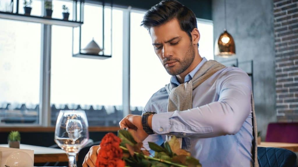 A man in a blue shirt looks at his wristwatch while sitting at a restaurant table.