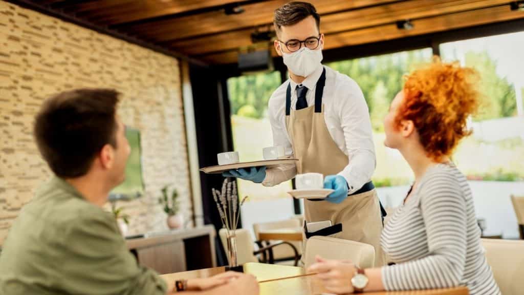 A masked waiter serves coffee on a tray to a man and woman seated at a table.