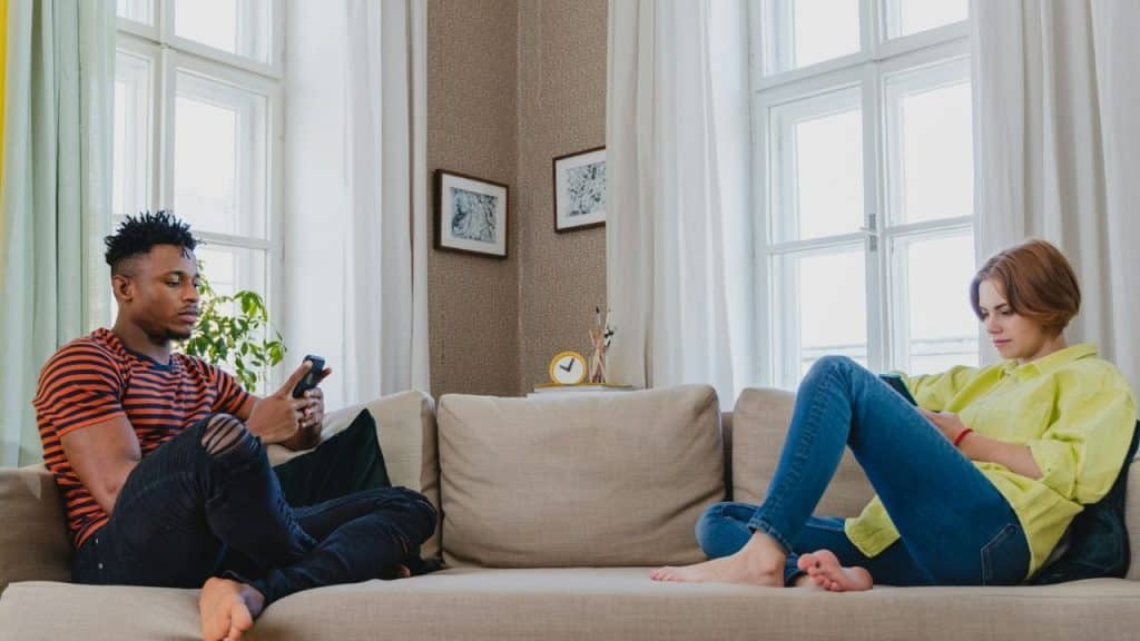 A man and woman sit on opposite ends of a couch while using their smartphones.