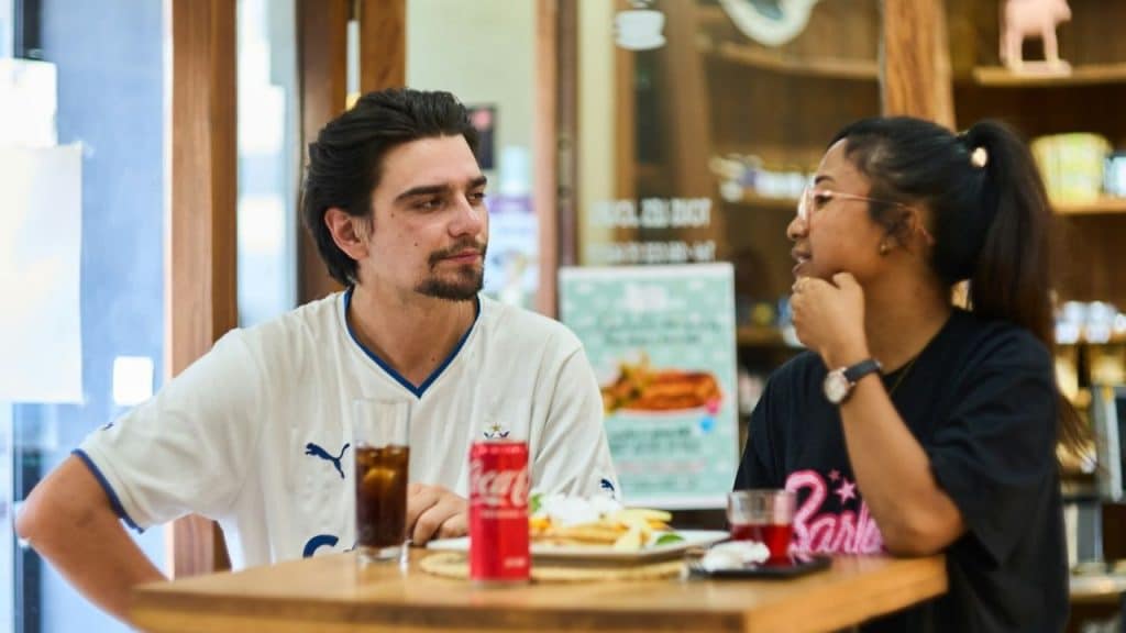 A man and woman sit at a wooden table with food and drinks while talking.