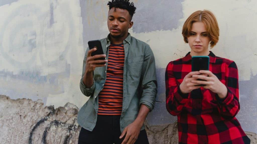 A man and woman lean against a wall while looking down at their separate smartphones.