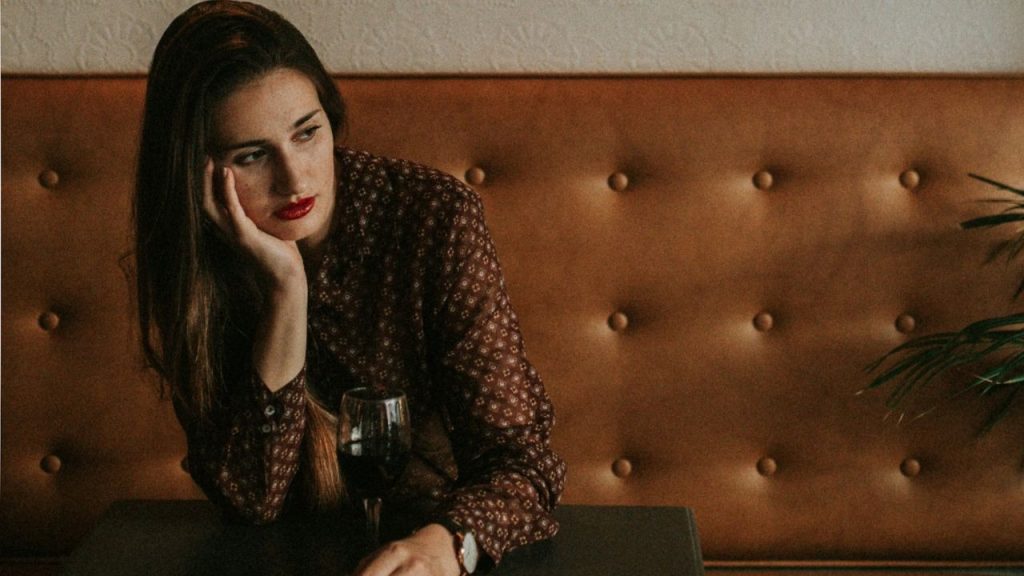 A woman with red lipstick leans on her hand next to a glass of wine.