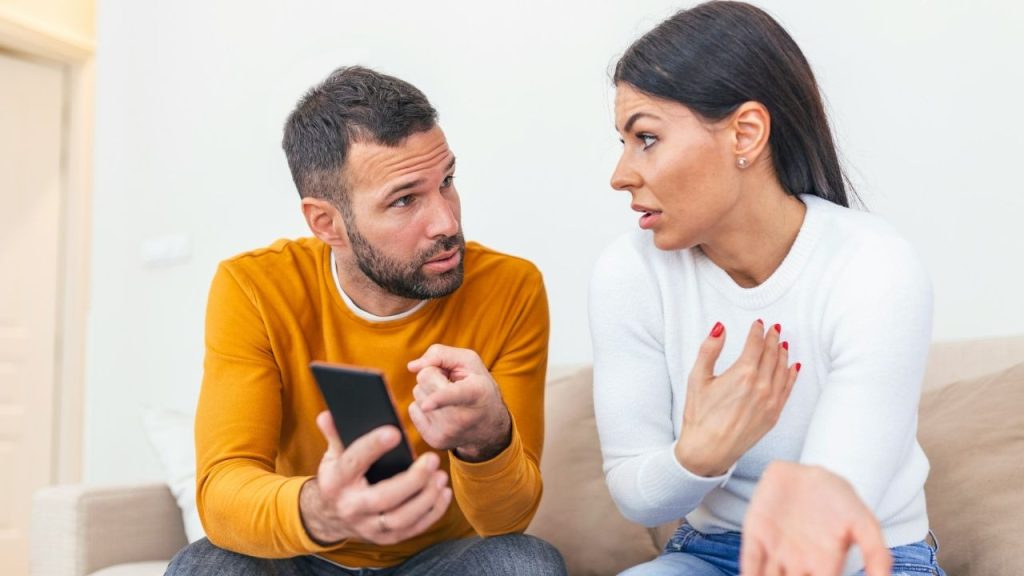 A man in a yellow shirt holds a phone while talking to a woman.