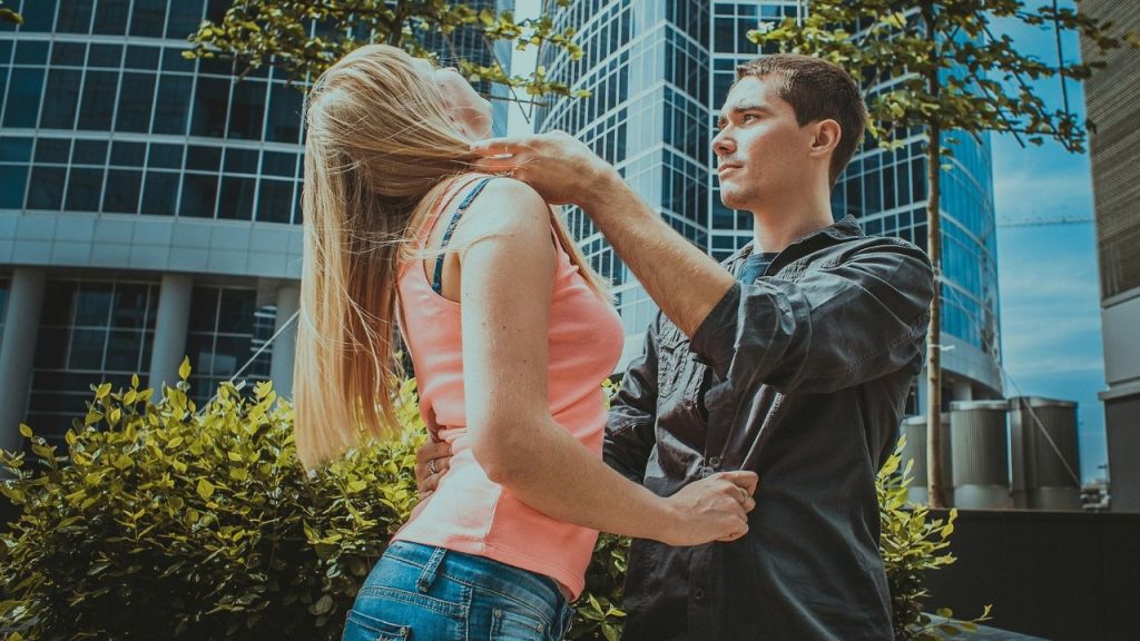 A man in a dark shirt touches a woman's neck in front of a building.