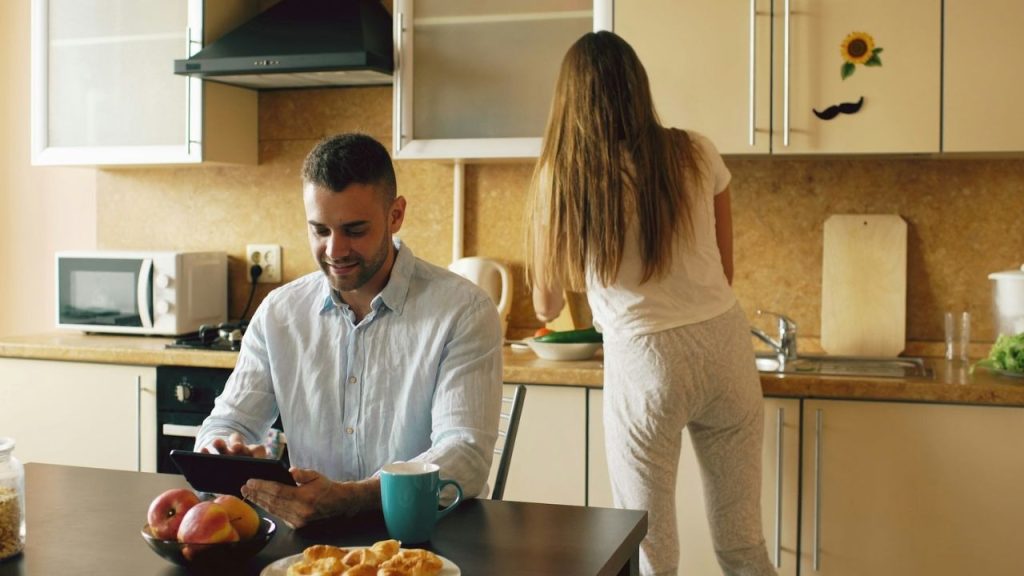 A man uses a tablet at a table while a woman works in the kitchen.