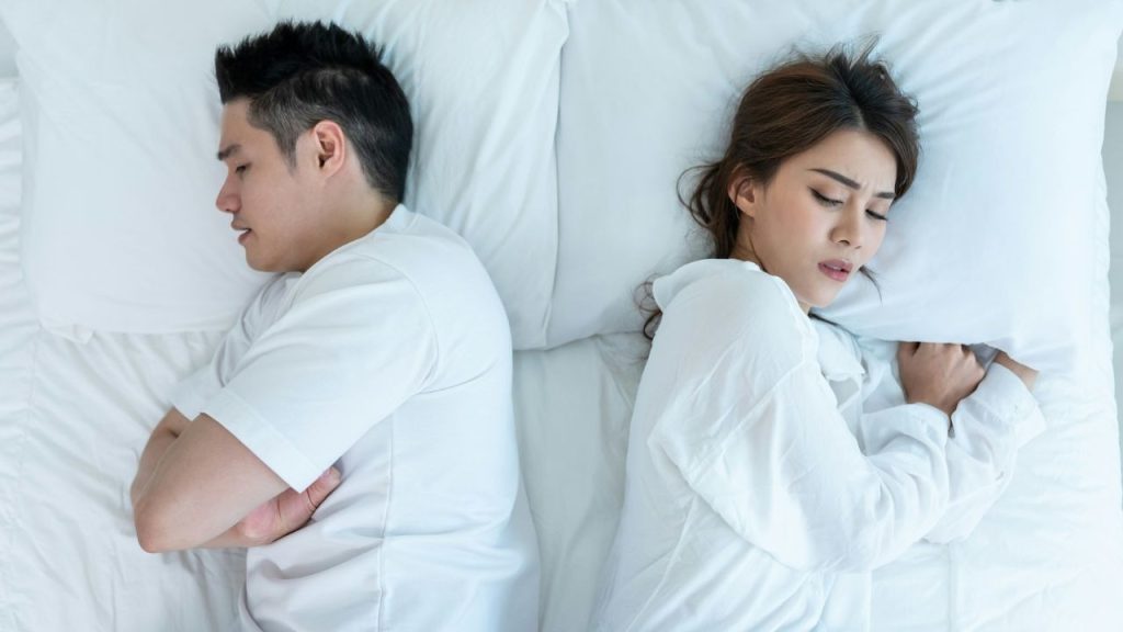 A man and woman lie back-to-back on a white bed with their eyes closed.