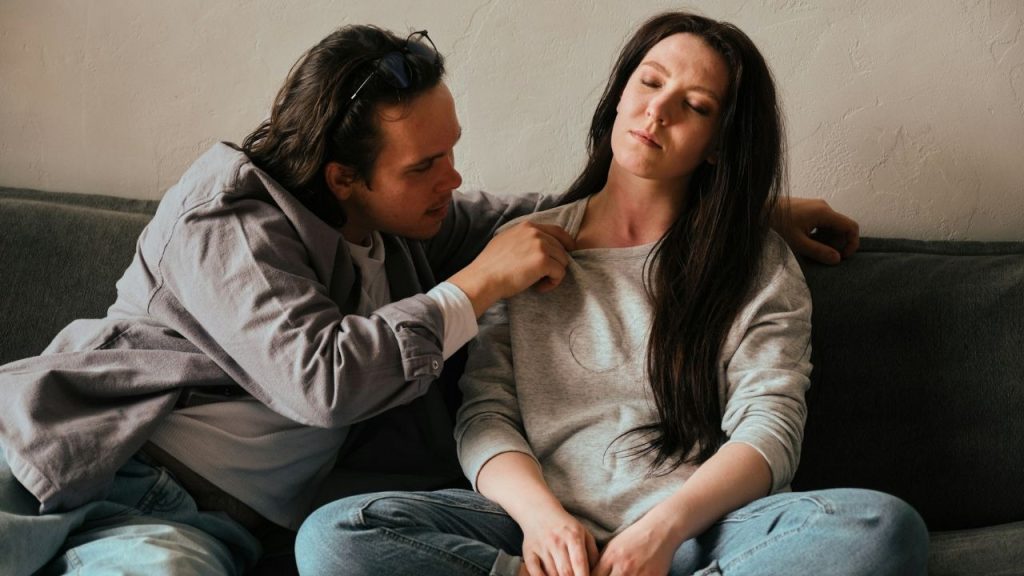 A man leans toward a woman sitting on a couch with her eyes tightly closed.