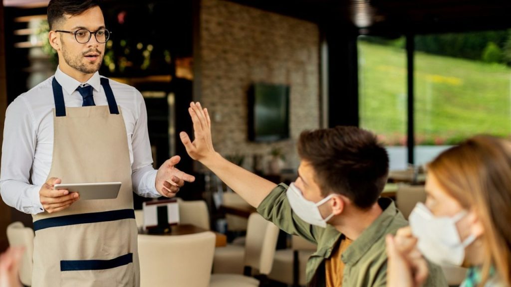 A man in a face mask holds up his hand to dismiss a restaurant server.