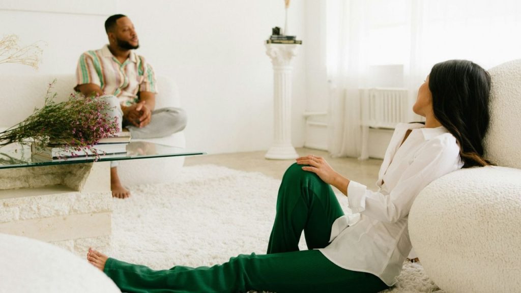 A woman sits on a white rug, leaning against a chair while looking toward a seated man.