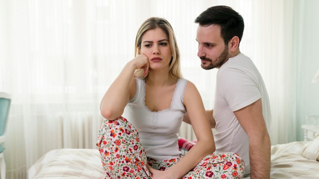 A blonde woman with a distressed expression is sitting cross-legged on a bed, while a man sits beside her looking at her with concern.