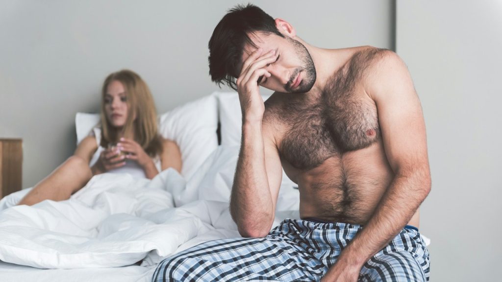A shirtless man sitting on a bed, looking distressed with his hand on his head, and a woman in the background.