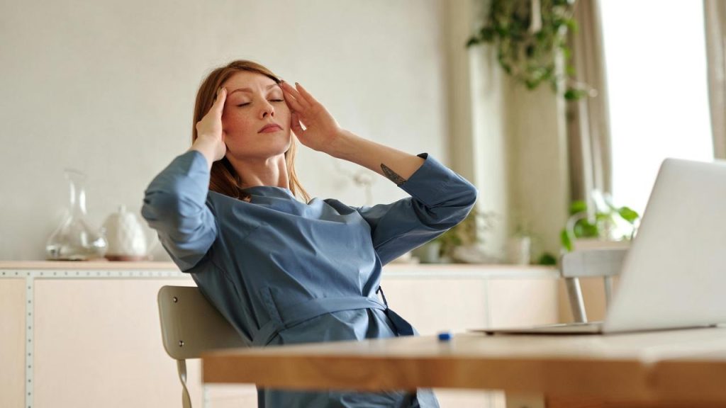 A woman sitting at a desk with a laptop, holding her temples as if stressed.