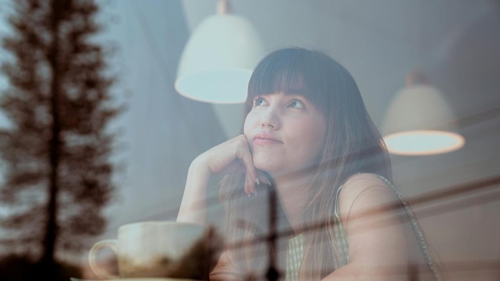 A woman gazing thoughtfully while sitting by a window with a cup in front of her.