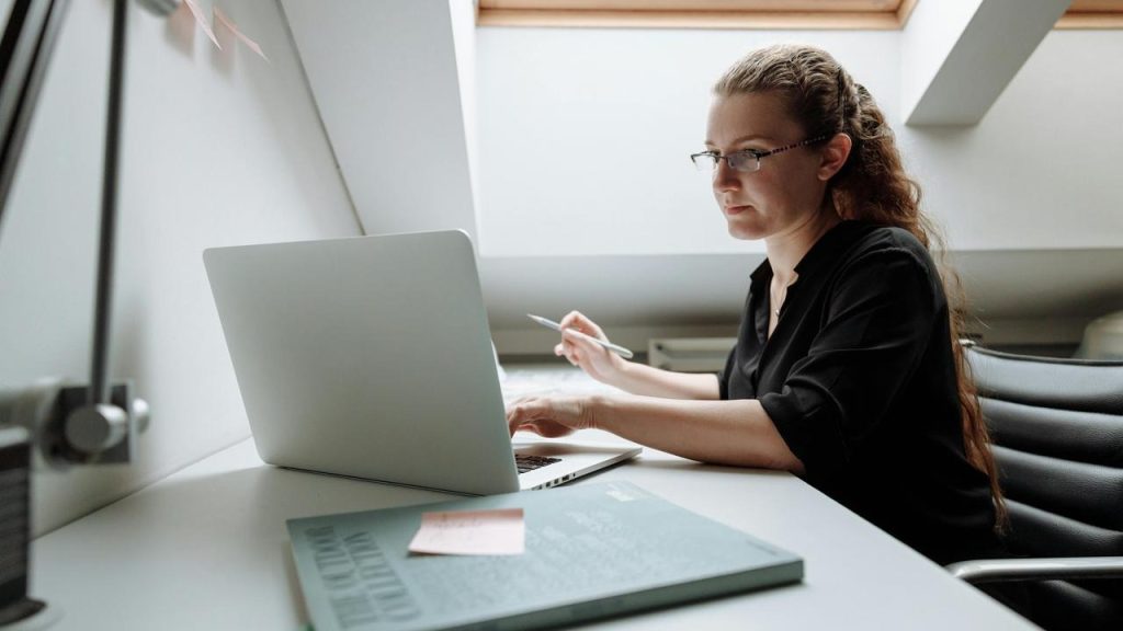 A woman working on a laptop while holding a pen at a desk.