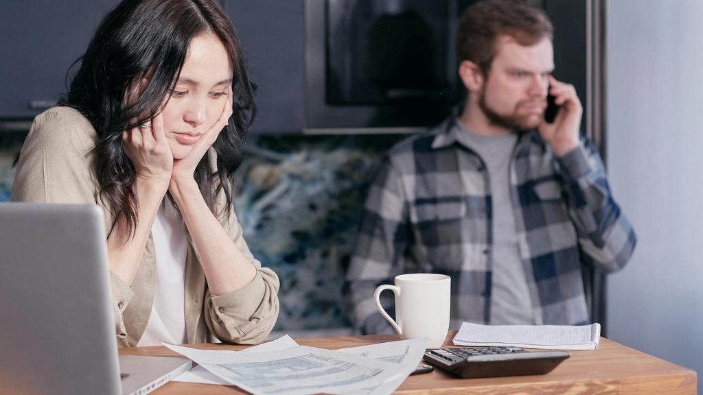 A worried woman reviewing papers beside a laptop while a man talks on the phone behind her.