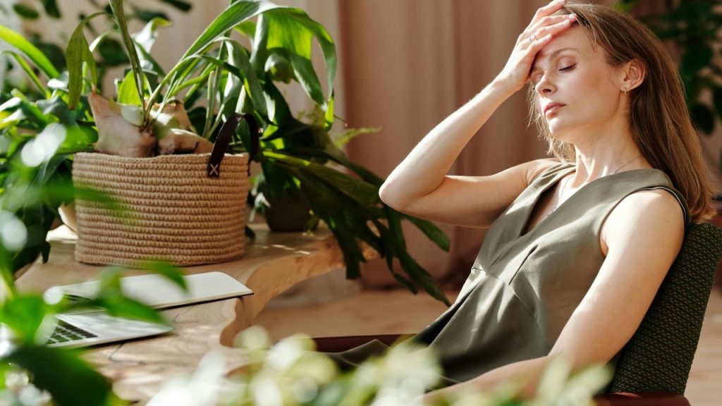 A woman sitting by a desk with plants, holding her forehead as if tired.