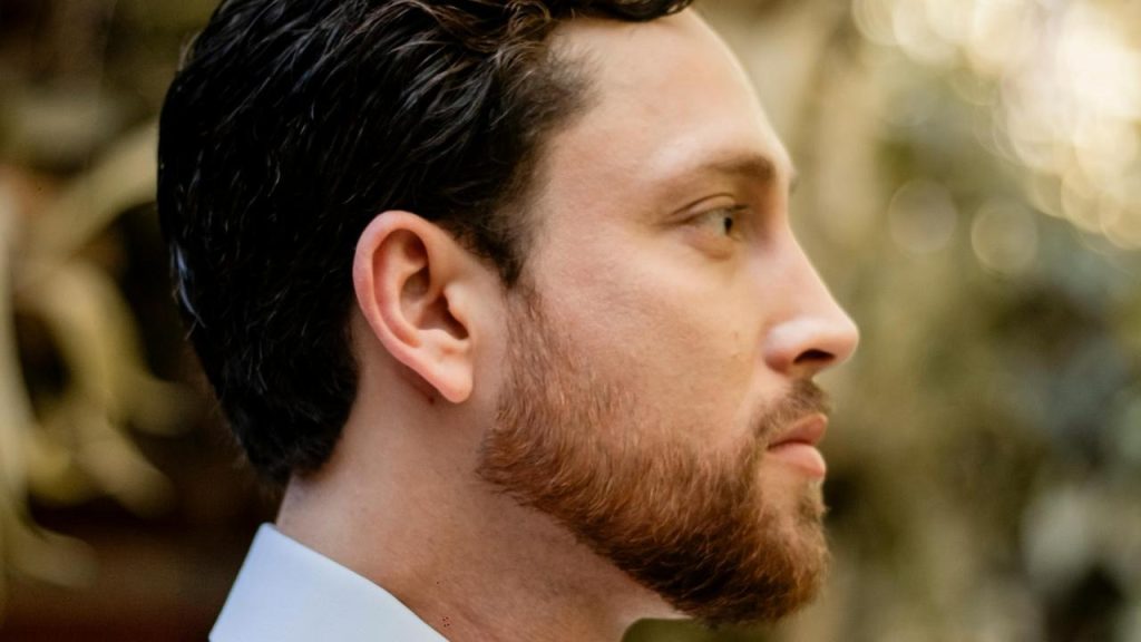 A side-profile of a bearded man with short dark hair.