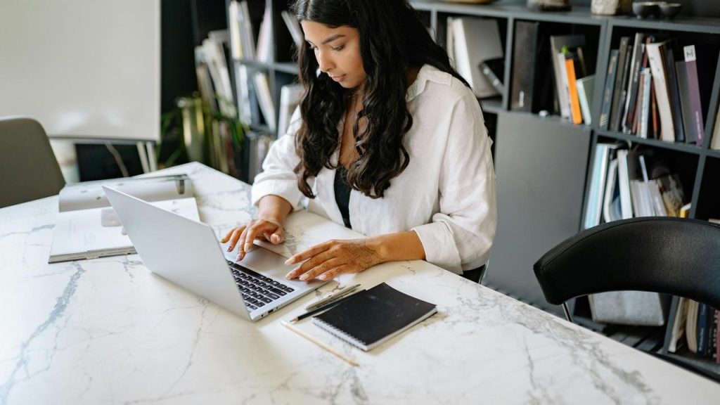 A woman working on a laptop at a desk with bookshelves in the background.