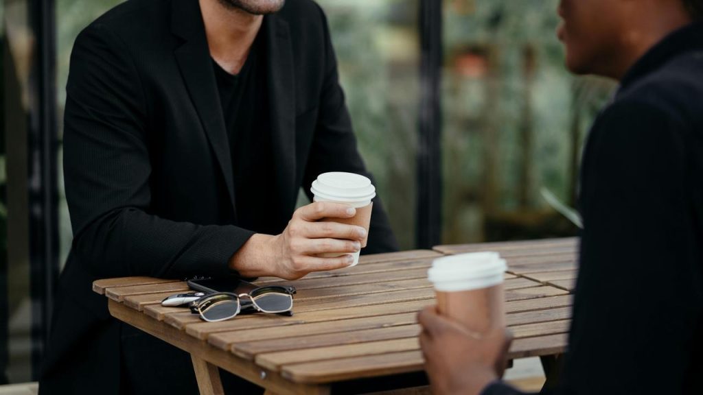 A pair of people sitting at a table holding takeaway coffee cups and talking.