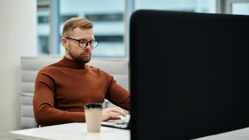 A man wearing glasses working on a laptop at an office desk.
