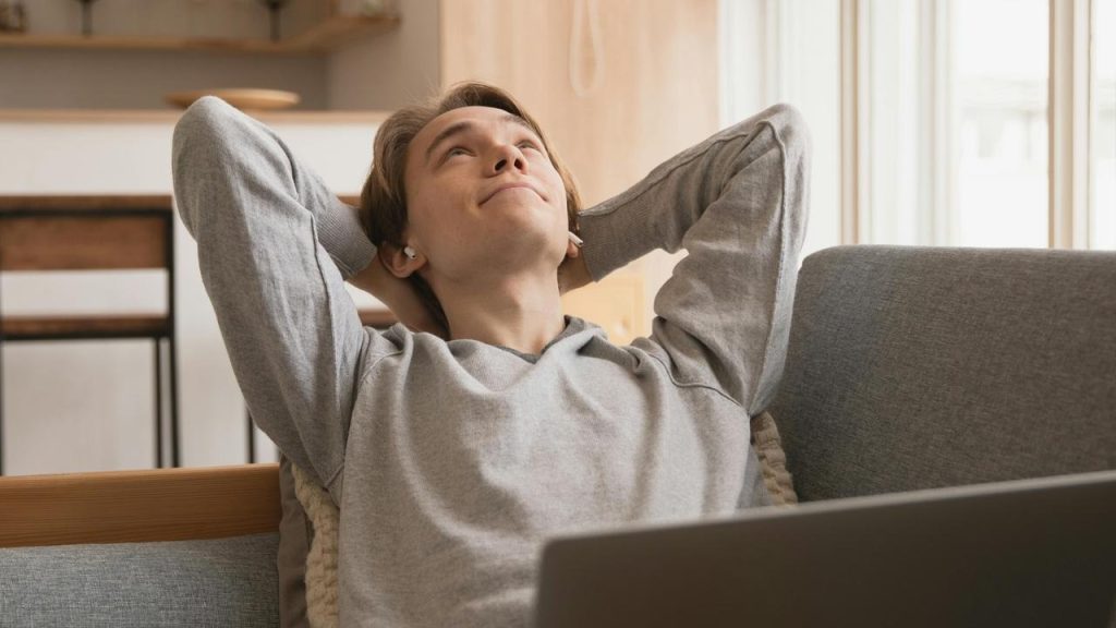 A relaxed man leaning back on a couch with a laptop nearby.