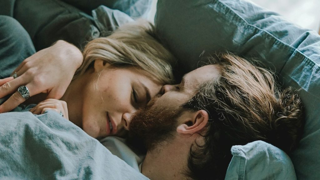 A bearded man kissing a woman's forehead while they lie together in a bed.