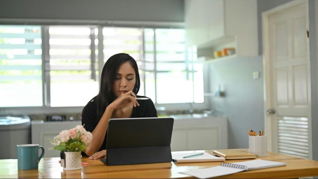 A woman working thoughtfully on a tablet at a kitchen table.