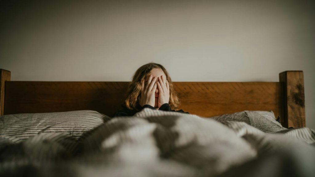 A person sitting in bed with hands covering their face, appearing distressed.