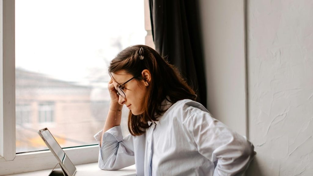 A woman wearing glasses, leaning by a window while looking at a tablet.
