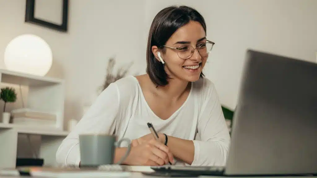 A smiling women wearing glasses and writing on a page while looking at a laptop’s screen.